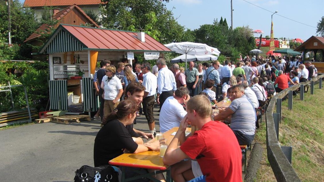 Eine lebendige Straßenszene mit vielen Menschen, die an Tischen sitzen und sich unterhalten. Im Hintergrund steht ein Verkaufsstand und zahlreiche Besucher sind unterwegs. | © Ga Weint Gehn Gemeinschaft