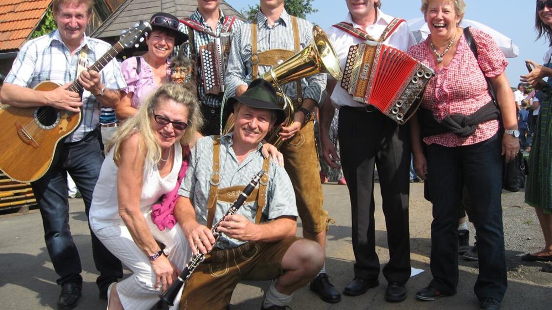 Eine fröhliche Gruppe von Menschen in traditioneller Kleidung, die Musikinstrumente spielen. Sie lächeln und posieren für ein Foto an einem sonnigen Tag. | © Ga Weint Gehn Gemeinschaft