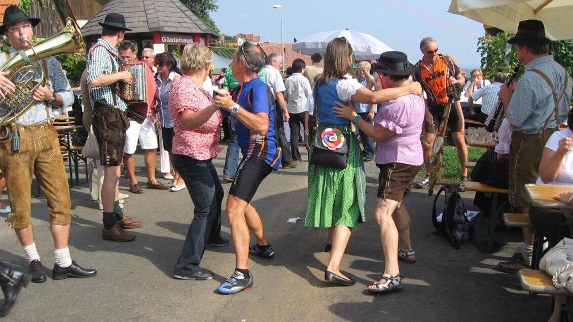 Eine fröhliche Feier auf der Straße mit Leuten in traditioneller Tracht. Man sieht tanzende Menschen und Musikanten, die eine festliche Stimmung schaffen. | © Ga Weint Gehn Gemeinschaft