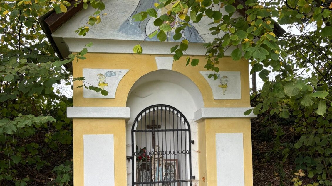 Gelb-weiße Kapelle bei Köflach mit Marienfigur hinter einem Eisengitter, religiösen Wandmalereien und umgebenden Bäumen. | © Kohlbacher