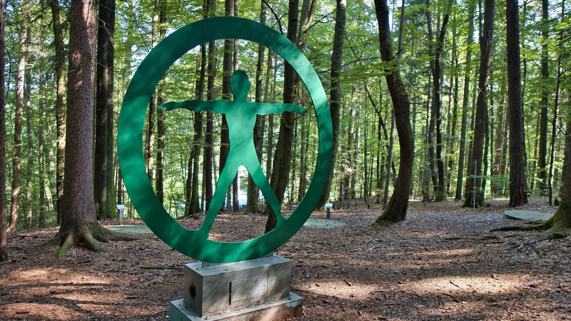 Grüne Metallskulptur Sebastian Reloaded mit stilisierter menschlicher Figur in einem Kreis, auf einem Sockel im Wald. | © Weges