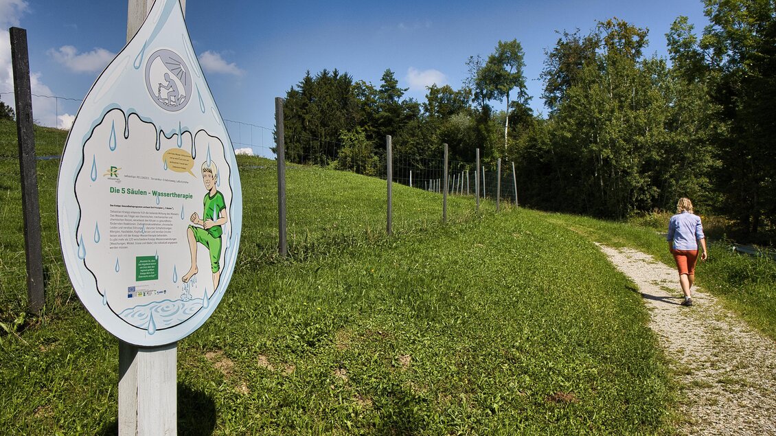 Wanderin auf einem Kiesweg in Laßnitzhöhe, vorbei an einer tropfenförmigen Infotafel zur 5-Säulen-Wassertherapie. | © Weges