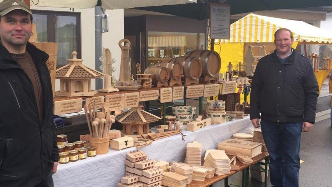 Zwei Männer stehen an einem Marktstand mit Holzprodukten. Der Stand zeigt verschiedene handgefertigte Holzartikel und Honiggläser. | © Marktgemeinde Gnas