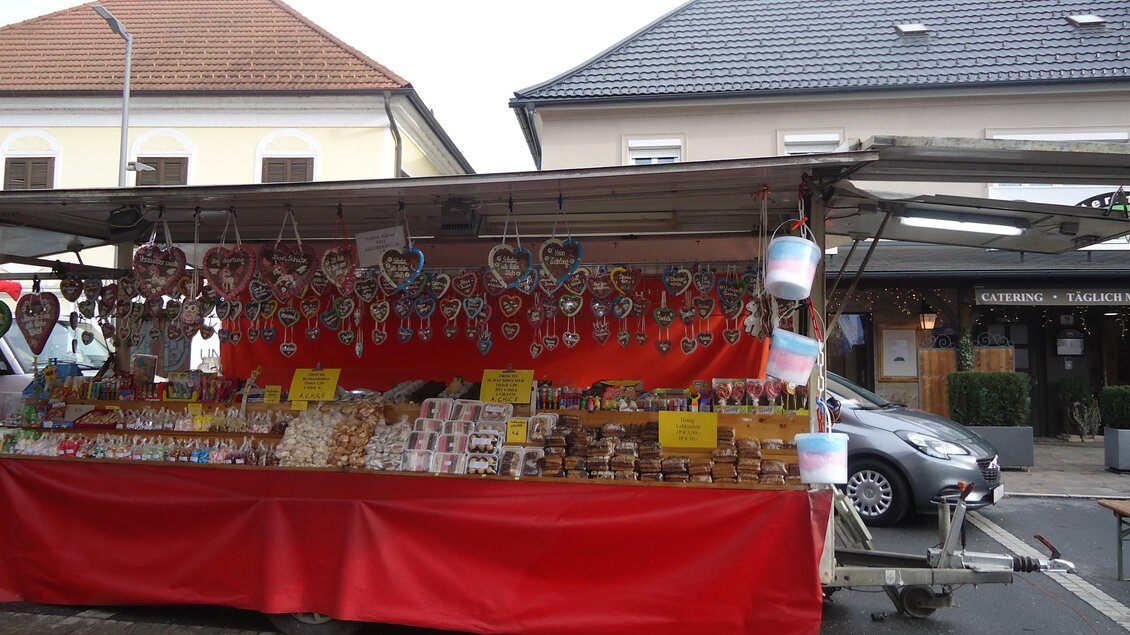 Ein Marktstand mit roten Tüchern, der eine Vielzahl von Süßigkeiten und Gebäck anbietet. Über dem Stand hängen bunte Dekorationen und Süßigkeiten. | © Marktgemeinde Gnas