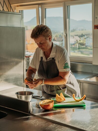 A chef is peeling fresh fruit in a modern kitchen. In the background, mountains and a clear sky can be seen. | © studio draussen