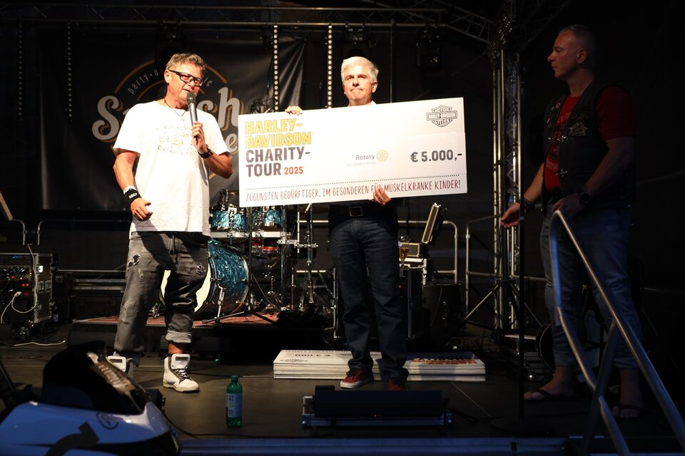 Drei Männer stehen auf einer Bühne und halten einen großen Scheck über 5.000 Euro. Im Hintergrund sind Musikinstrumente und ein Schild mit dem Schriftzug "Charity Tour 2023" zu sehen. | © Zehnerhaus