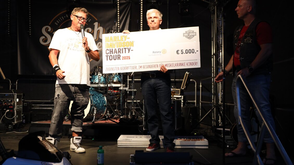 Drei Männer stehen auf einer Bühne und halten einen großen Scheck über 5.000 Euro. Im Hintergrund sind Musikinstrumente und ein Schild mit dem Schriftzug "Charity Tour 2023" zu sehen. | © Zehnerhaus