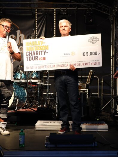 Drei Männer stehen auf einer Bühne und halten einen großen Scheck über 5.000 Euro. Im Hintergrund sind Musikinstrumente und ein Schild mit dem Schriftzug "Charity Tour 2023" zu sehen. | © Zehnerhaus