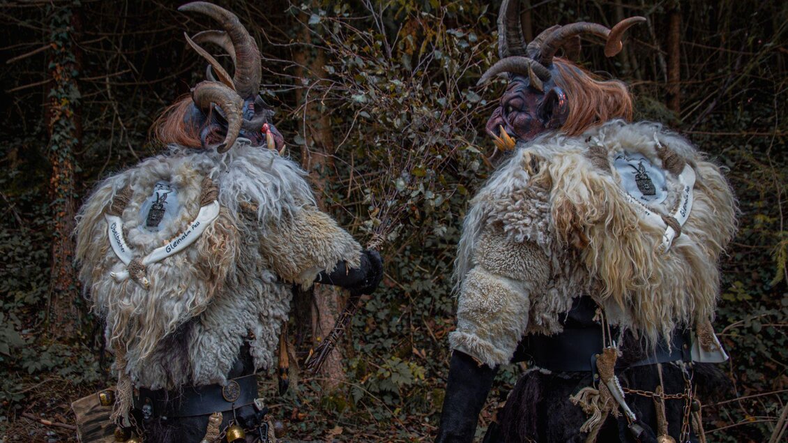 Zwei Menschen in traditionellen Kostümen mit Hörnern und Fellmänteln stehen in einem Wald. Der Hintergrund ist mit grünen Pflanzen umgeben. | © Übelbacher Gleinalm Pass