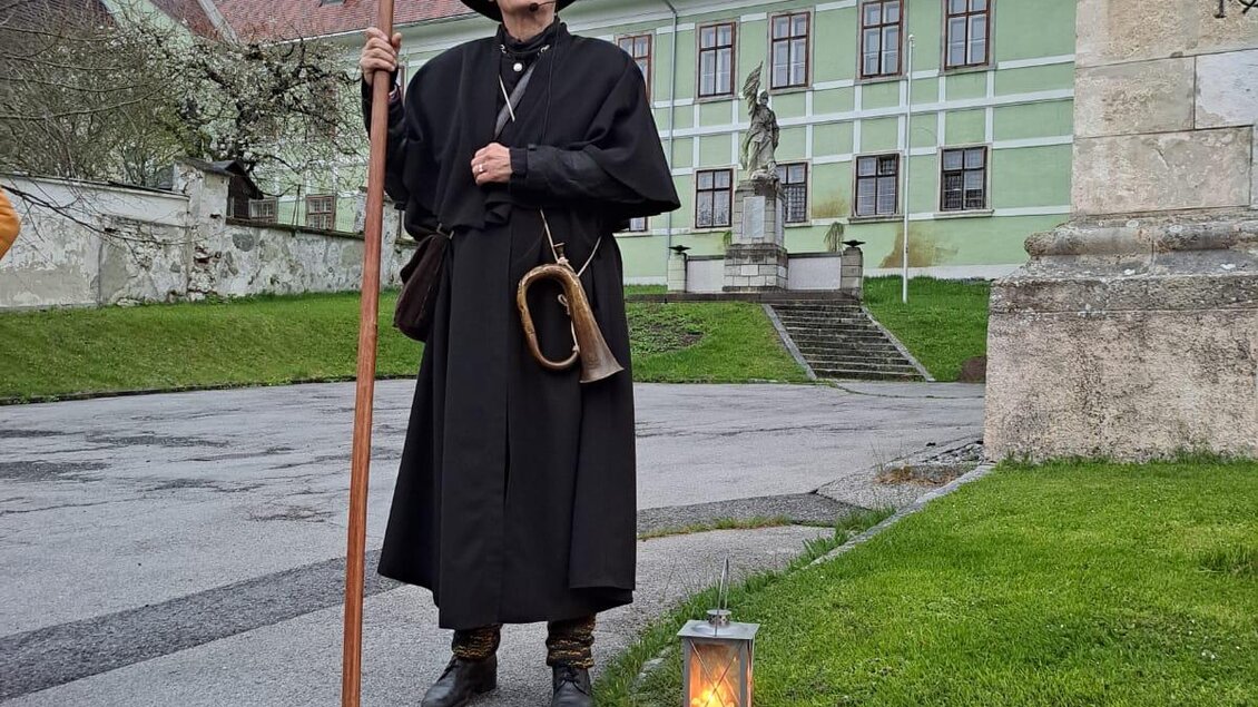 Ein historisch gekleideter Mann steht mit einer Lanze in der Hand an einer Straßenkreuzung. Im Hintergrund ist ein Gebäude mit grüner Fassade zu sehen. | © © Kurverwaltung Aflenz
