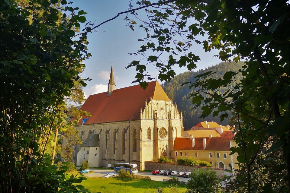 Eine beeindruckende Kirche mit rotem Dach und hohen Fenstern, umgeben von Bäumen. Im Vordergrund sieht man eine grüne Wiese und einige Gebäude. | © Andreas Steininger