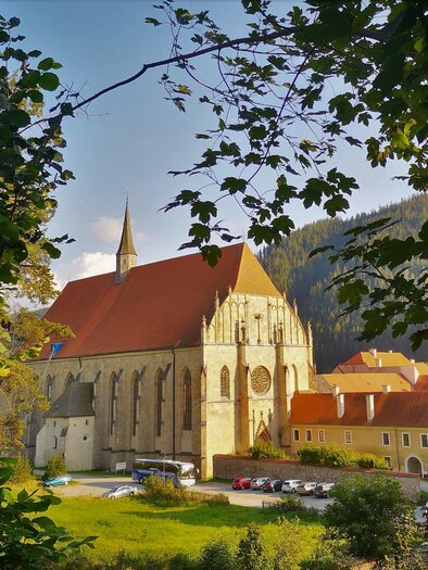 Eine beeindruckende Kirche mit rotem Dach und hohen Fenstern, umgeben von Bäumen. Im Vordergrund sieht man eine grüne Wiese und einige Gebäude. | © Andreas Steininger