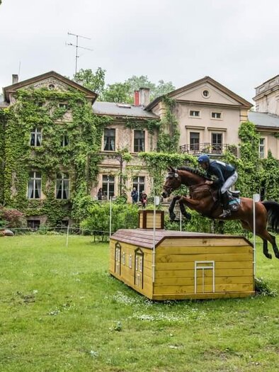 Ein Reiter springt mit seinem Pferd über ein Hindernis vor einem alten, von Efeu bewachsenen Gebäude. Die Szene spielt in einer grünen Landschaft mit Bäumen im Hintergrund. | © Lisa Held-Dunst