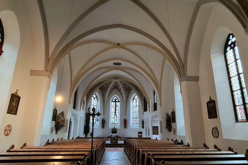 Eine helle Kirche mit hohen Gewölben und bunten Fenstern. Reihen von Holzbanken verlaufen bis zum Altar. | © Stephanie Bor