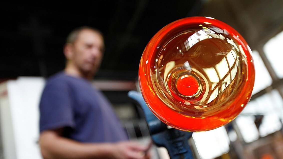 Ein Glasbläser arbeitet mit heißem, flüssigem Glas. Im Vordergrund ist ein leuchtend rotes Glasobjekt zu sehen. | © Enrico Caracciolo