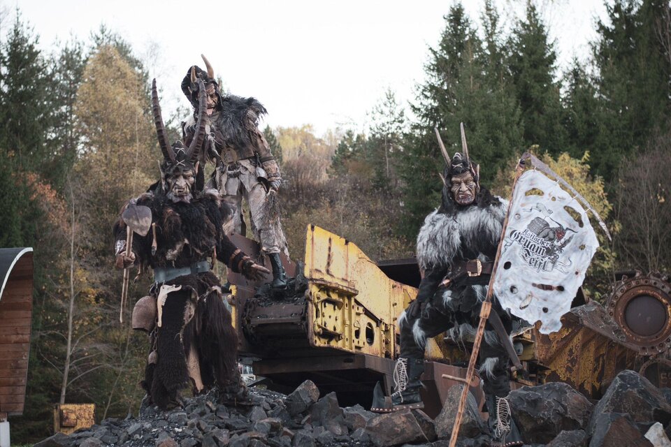 Drei maskierte Figuren in beeindruckenden Kostümen stehen auf einem Steinhaufen. Im Hintergrund sind baumbewachsene Hügel und eine alte Maschine sichtbar. | © Jessica Schutting