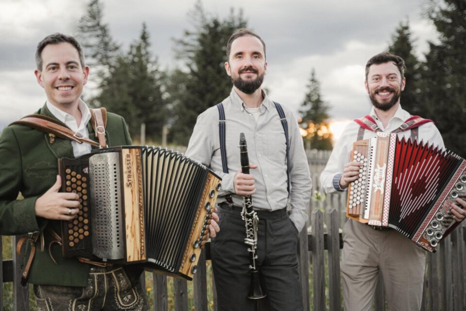Drei Musiker mit Harmonika und Klarinette stehen vor Holzzaun auf Almwiese in der Steiermark bei Sonnenuntergang. | © Reithofer Media