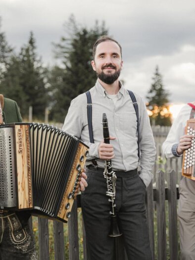 Drei Musiker mit Harmonika und Klarinette stehen vor Holzzaun auf Almwiese in der Steiermark bei Sonnenuntergang. | © Reithofer Media