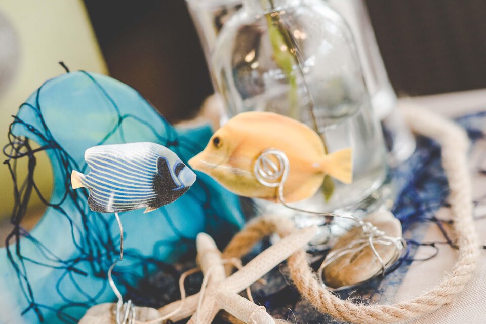 Ein dekoratives Arrangement mit zwei bunten Fischen und einem Glas mit Wasser. Das alles ist umgeben von einem Netz und einem Seil. | © Garten Hotel Ochensberger