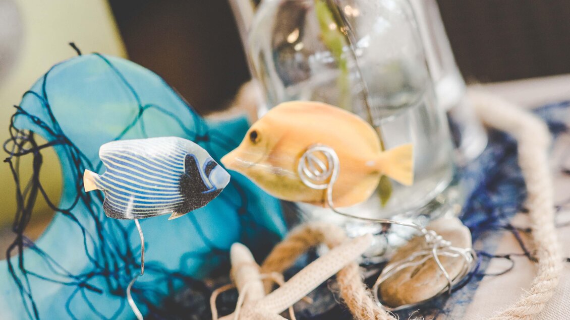Ein dekoratives Arrangement mit zwei bunten Fischen und einem Glas mit Wasser. Das alles ist umgeben von einem Netz und einem Seil. | © Garten Hotel Ochensberger