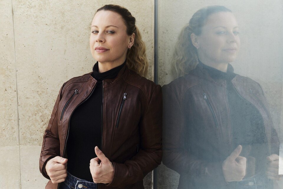 Katharina Straßer in brauner Lederjacke und schwarzem Oberteil lehnt an einer Glaswand, in der sich ihr Spiegelbild zeigt. | © Hilde van Mas
