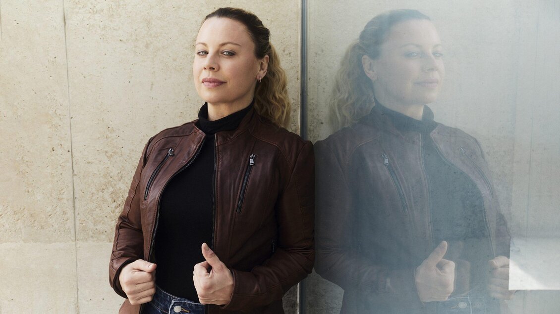 Katharina Straßer in brauner Lederjacke und schwarzem Oberteil lehnt an einer Glaswand, in der sich ihr Spiegelbild zeigt. | © Hilde van Mas