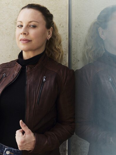 Katharina Straßer in brauner Lederjacke und schwarzem Oberteil lehnt an einer Glaswand, in der sich ihr Spiegelbild zeigt. | © Hilde van Mas