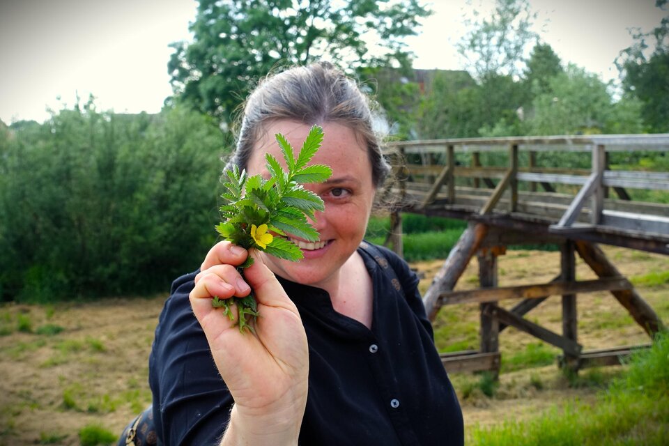 Eine Frau hält ein kleines Bündel frischer Pflanzen in der Hand. Im Hintergrund sind Bäume und eine Holzbrücke zu sehen. | © Martina Schmidt