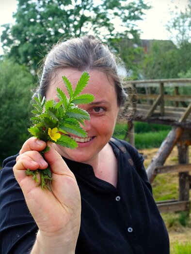 Eine Frau hält ein kleines Bündel frischer Pflanzen in der Hand. Im Hintergrund sind Bäume und eine Holzbrücke zu sehen. | © Martina Schmidt