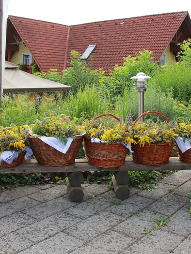 Eine Holzbank mit mehreren Korbgefäßen, die mit gelben Blumen gefüllt sind. Im Hintergrund ist ein grüner Garten und ein rotes Dachhaus sichtbar. | © Notburga Meister