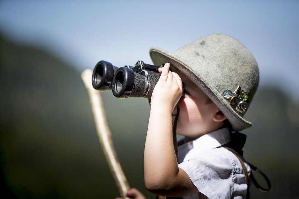 Ein kleines Kind mit einem faszinierenden Hut schaut durch ein Fernglas. Es hält einen Stock in der Hand und blickt neugierig in die Ferne. | © Tom Lamm