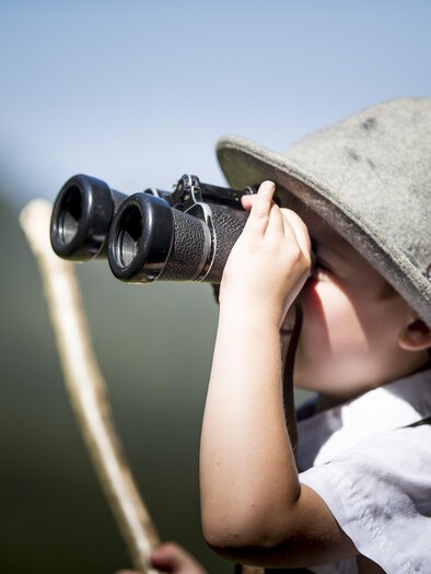 A small child with a fascinating hat is looking through binoculars. It is holding a stick in its hand and curiously gazing into the distance. | © Tom Lamm