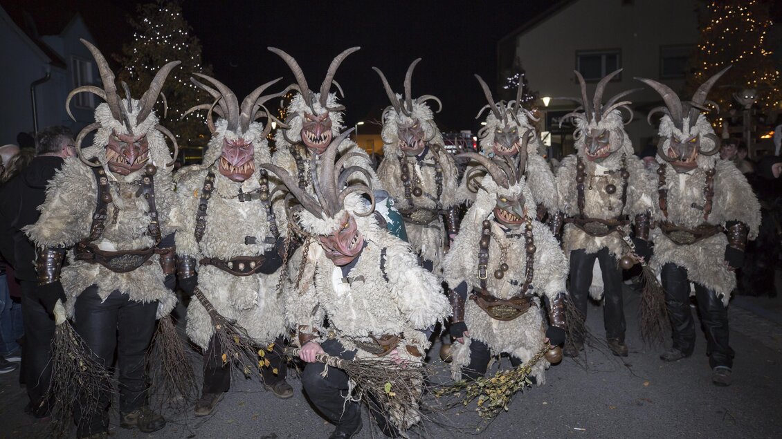 Eine Gruppe von verkleideten Personen in traditionellen Kostümen mit Hörnern und Fellen. Sie stehen in einer festlichen Umgebung bei Nacht. | © Wabi