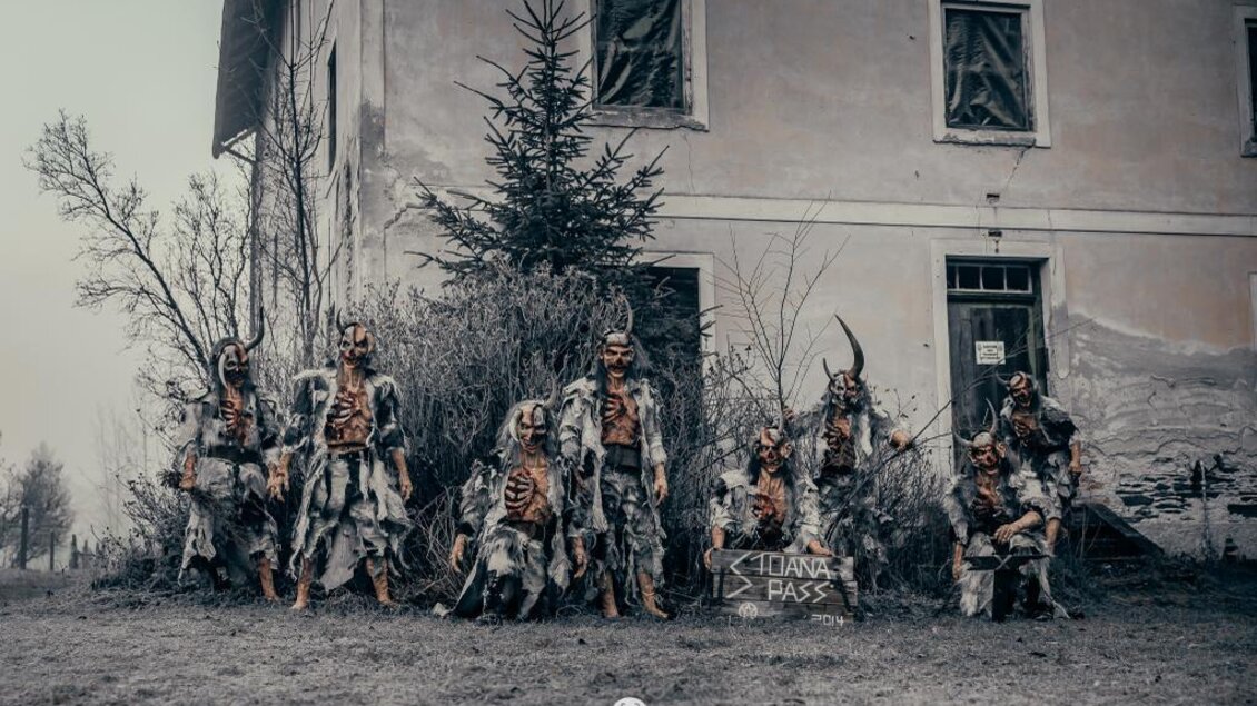 Eine gruselige Szene mit mehreren Figuren in unheimlichen Kostümen vor einem alten Gebäude. Die Atmosphäre ist düster und unheimlich, ergänzt durch eine verwilderte Umgebung. | © Stoana Pass
