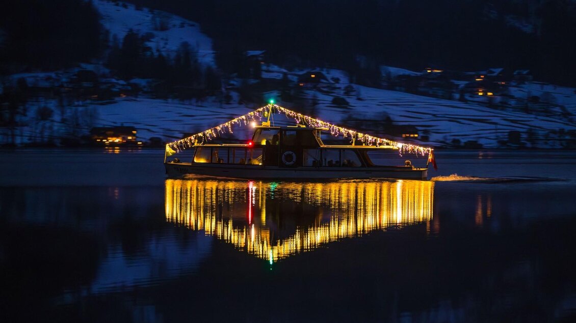 Die beleuchtete MS Traun – ein Schiff aus der Flotte der Schifffahrt Grundlsee – fährt im Dunkeln über den ruhigen Grundlsee. Im Hintergrund sind schneebedeckte Berge und einige hell erleuchtete Häuser sichtbar. | © Schifffahrt Grundlsee