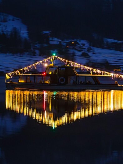 Die beleuchtete MS Traun – ein Schiff aus der Flotte der Schifffahrt Grundlsee – fährt im Dunkeln über den ruhigen Grundlsee. Im Hintergrund sind schneebedeckte Berge und einige hell erleuchtete Häuser sichtbar. | © Schifffahrt Grundlsee