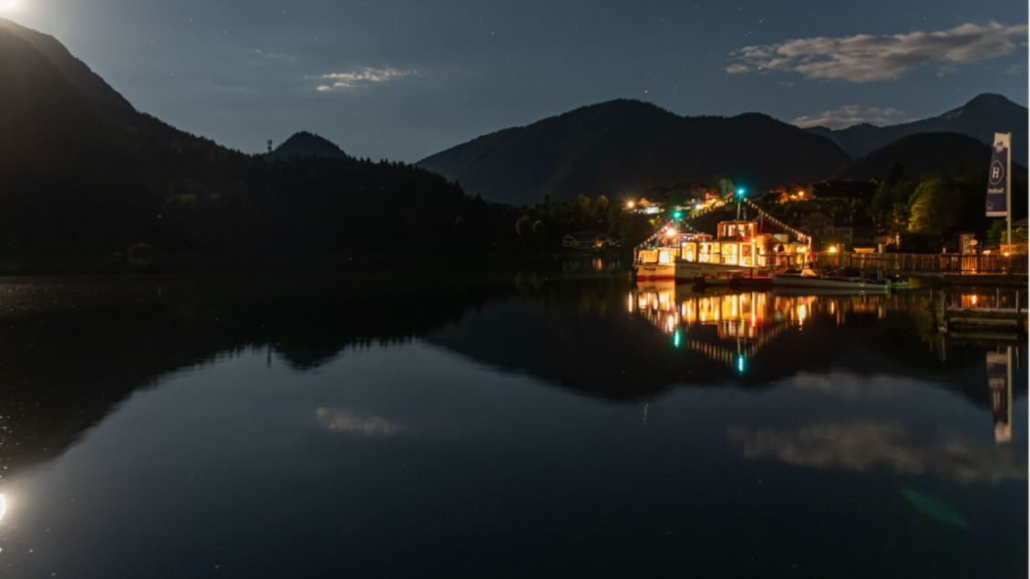 Ein ruhiger See bei Nacht mit spiegelndem Wasser. Im Hintergrund sind Berge und eine freundlich beleuchtete Hütte zu sehen. | © Schifffahrt Grundlsee