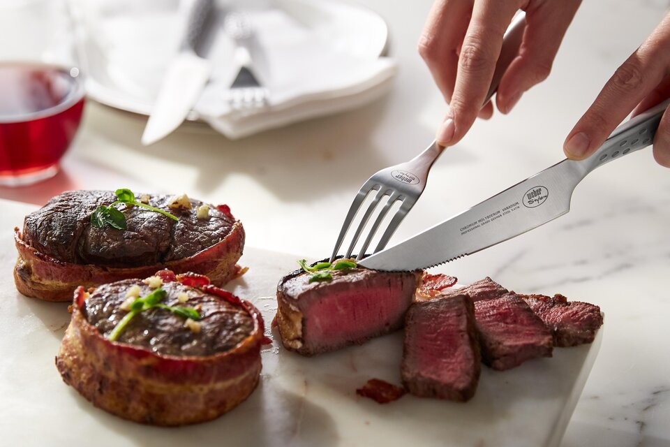 Ein saftiges Stück Fleisch wird auf einem Schneidebrett aufgeschnitten. Im Hintergrund ist ein Glas Wein und Geschirr zu sehen. | © Weber Grill