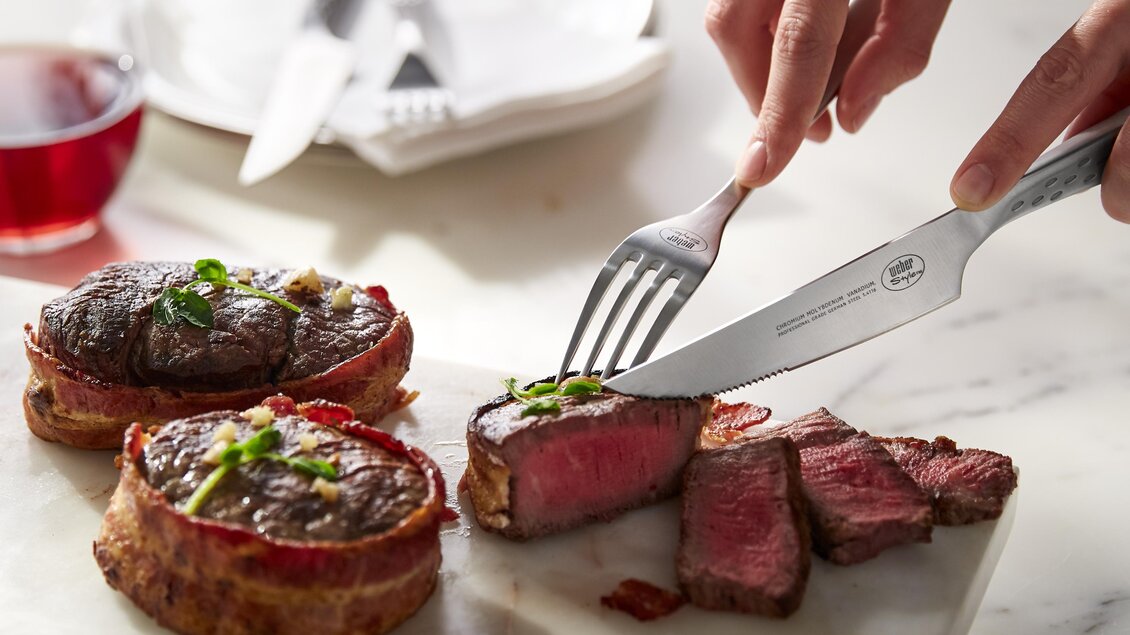 Ein saftiges Stück Fleisch wird auf einem Schneidebrett aufgeschnitten. Im Hintergrund ist ein Glas Wein und Geschirr zu sehen. | © Weber Grill