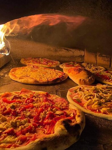 Mehrere belegte Pizzen backen neben offenem Feuer in einem traditionellen Holzofen. | © pizzerija-pod-gradom