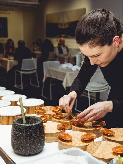 Chef Jaimy Reisinger artfully arranges small dishes on wooden and stone plates in the kitchen and prepares a culinary experience with great attention to detail. | © Pixelcoma-Cliff Kapatais