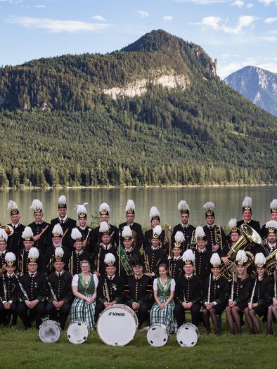 The Salinenmusikkapelle Altaussee is a large brass band. The members of the Salinenmusikkapelle stand on the shore of Lake Altaussee, which is surrounded by mountains. The musicians wear the traditional black uniform of the Saline Altaussee and hats. | © Erich Kainzinger