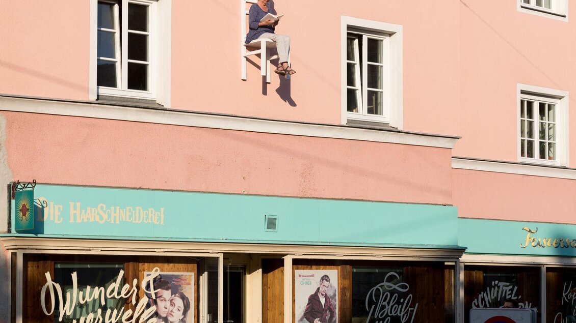 Ein Mann sitzt in einem Fenster auf einer Sitzbank und liest. Die Fassade des Gebäudes ist in sanften Rosa- und Blautönen gehalten. | © Graz Tourismus - Harry Schiffer