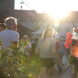 Ein Glas Getränk mit Früchten im Vordergrund und Menschen, die in der Abendsonne spazieren gehen, im Hintergrund. Die Szene vermittelt eine entspannte und fröhliche Atmosphäre. | © Thermen&Vulkanland