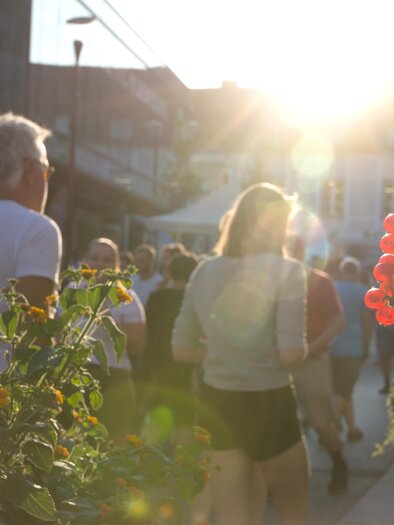 Ein Glas Getränk mit Früchten im Vordergrund und Menschen, die in der Abendsonne spazieren gehen, im Hintergrund. Die Szene vermittelt eine entspannte und fröhliche Atmosphäre. | © Thermen&Vulkanland