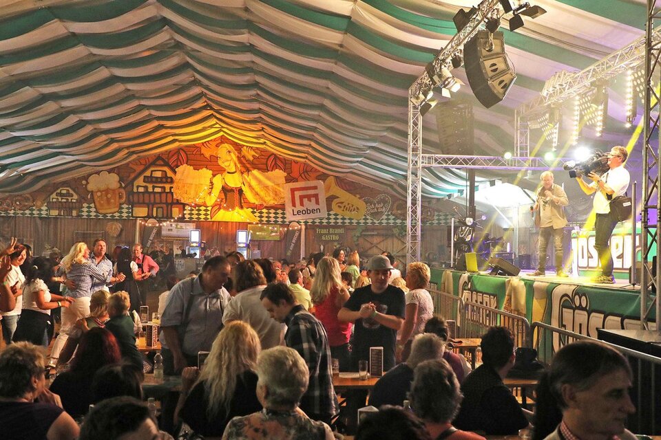 Eine festliche Veranstaltung in einem beheizten Zelt mit Menschen, die tanzen und feiern. Auf der Bühne treten Musiker auf, während die Stimmung lebhaft und fröhlich ist. | © Wiesenfest