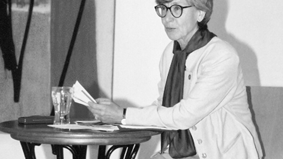 Hedwig Wingler liest bei einer Veranstaltung im Kunsthaus Köflach aus einem Buch und sitzt dabei an einem Tisch vor einer Präsentationsfläche. | © agathon koren