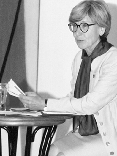 Hedwig Wingler liest bei einer Veranstaltung im Kunsthaus Köflach aus einem Buch und sitzt dabei an einem Tisch vor einer Präsentationsfläche. | © agathon koren