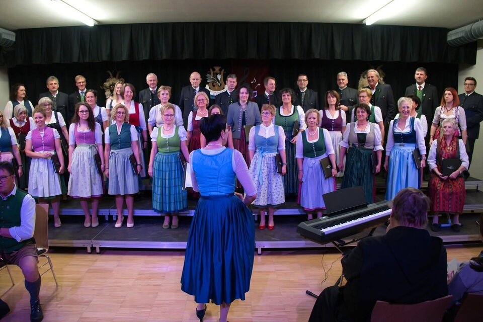 Eine große Gruppe von Sängern steht auf der Bühne in traditioneller Tracht und singt gemeinsam. Im Vordergrund befindet sich ein Mann am Klavier, während das Publikum zuschaut. | © echtzeit tv