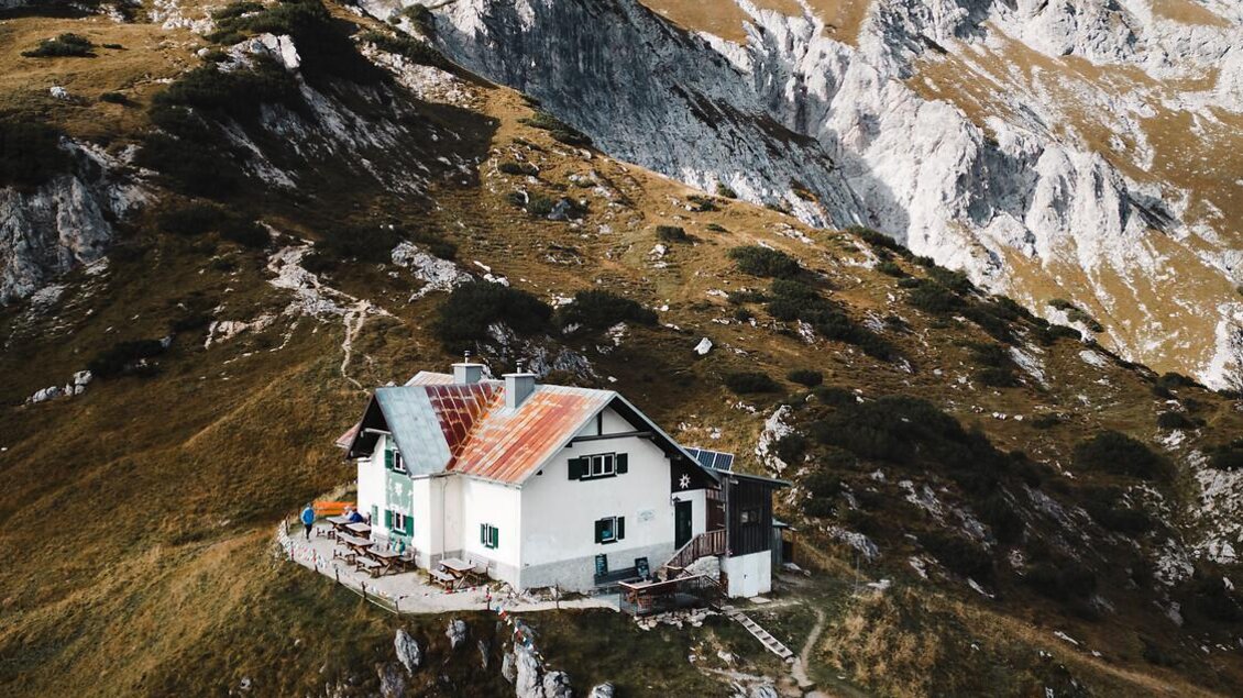 Eine einsame Berghütte auf einem Hügel, umgeben von grünen Wiesen und Felsen. Im Hintergrund erheben sich majestätische Berge unter einem klaren Himmel. | © TV Gesäuse